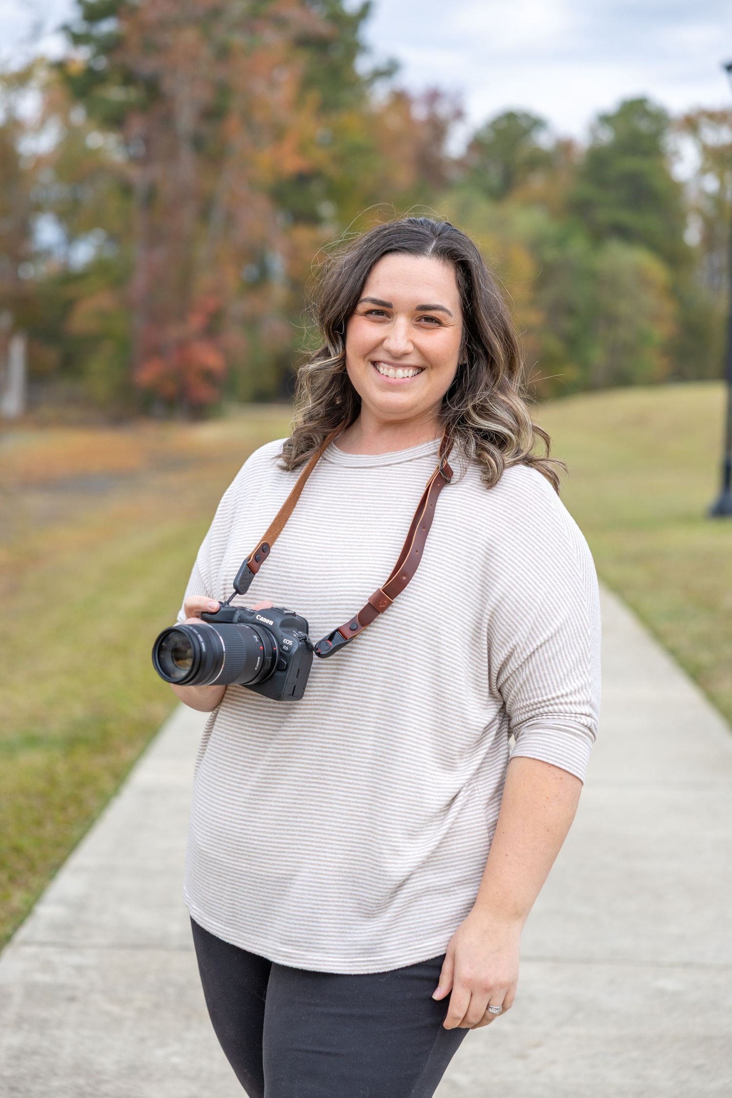Photographer in white sweater with camera strap smiles during outdoor portrait session.