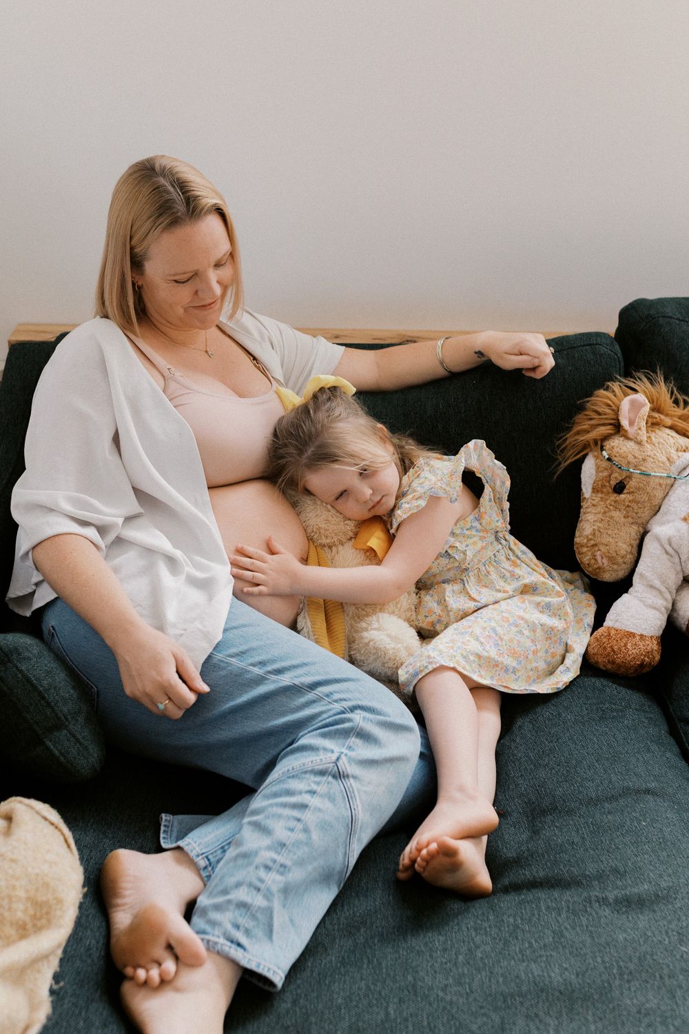 Mother and child resting together on dark gray couch with stuffed animals and pillows.