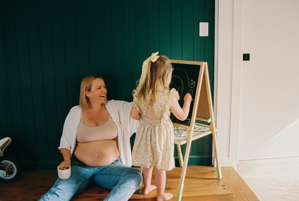 Two people in a room with a green wall and an easel chalkboard, one sitting and one standing drawing.