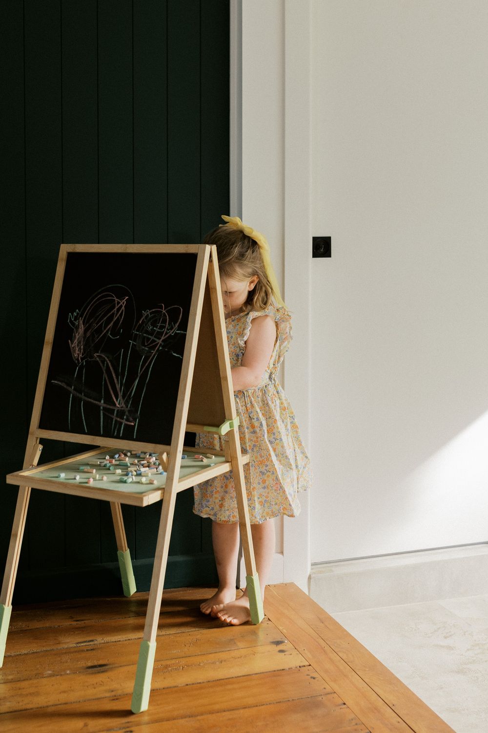 A child stands at a small wooden easel with mint green legs drawing on a chalkboard in a minimalist room.