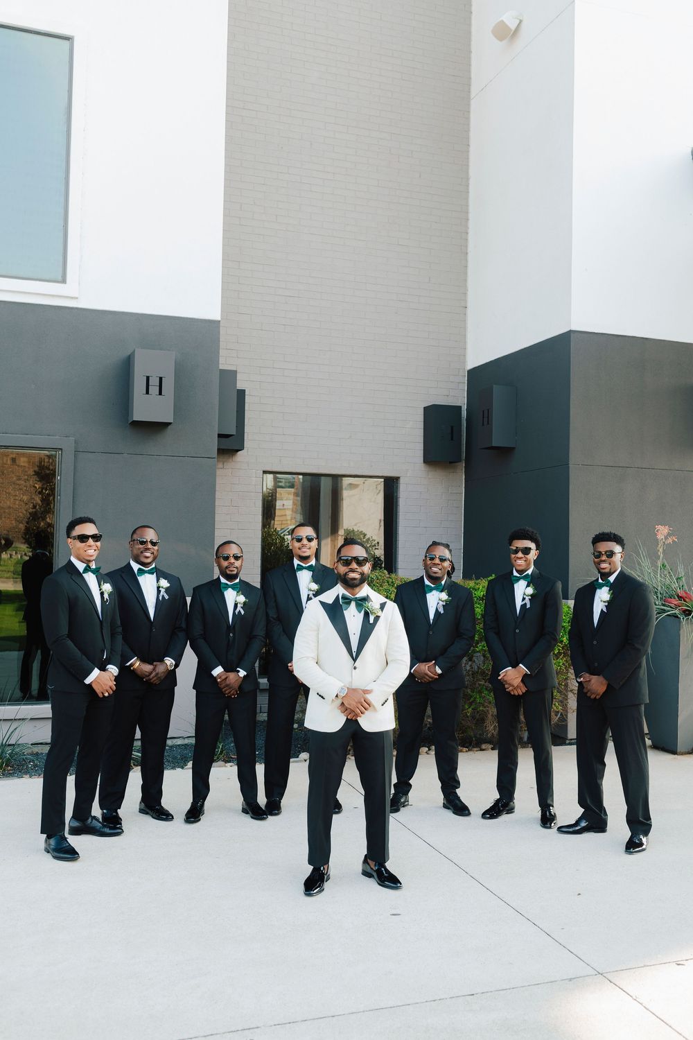 A group of groomsmen in black suits and sunglasses pose together against a white wall for a stylish wedding photo.