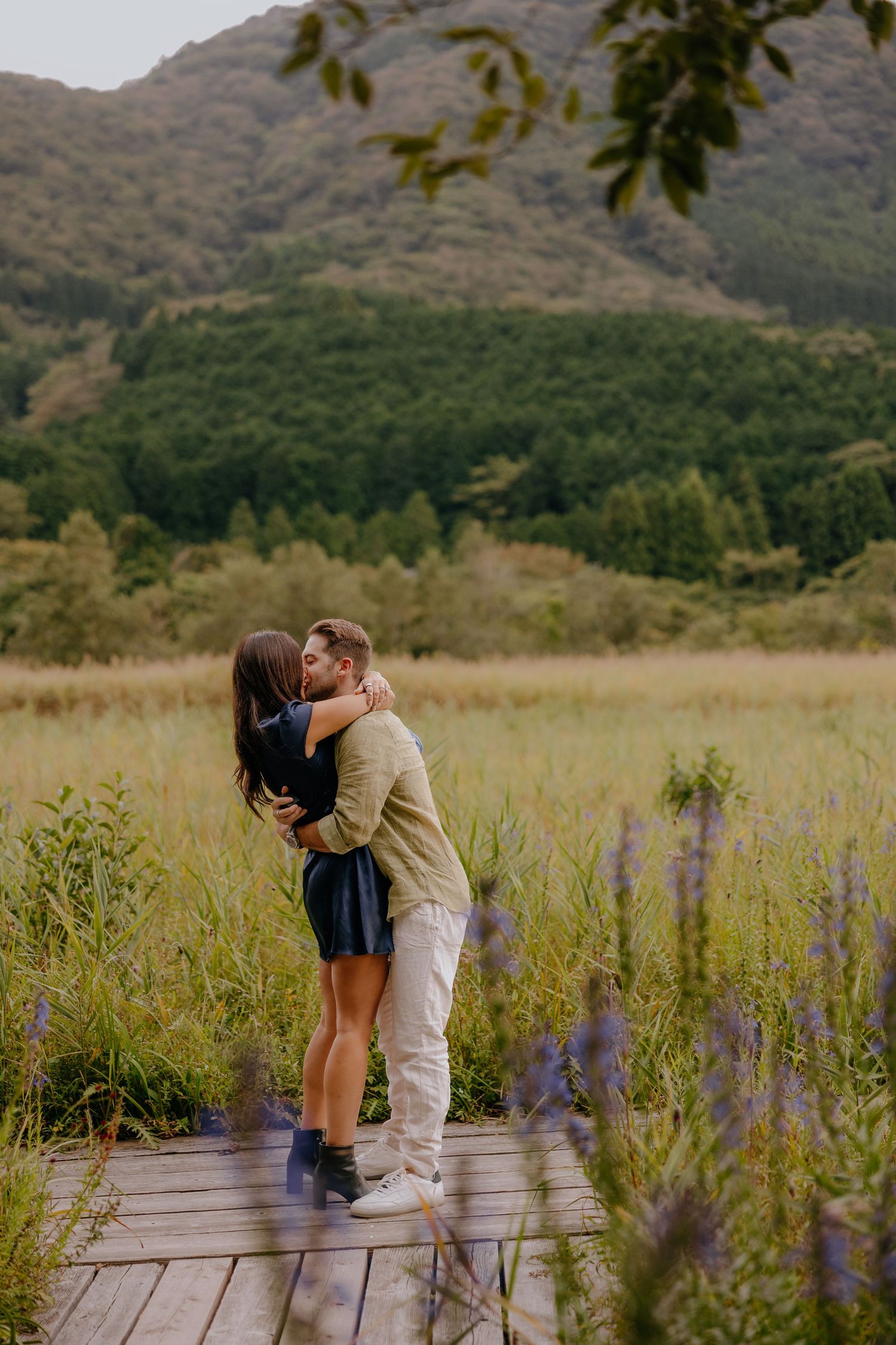 A romantic couple shares a tender moment on a wooden boardwalk surrounded by purple wildflowers and mountain scenery at Hakone Japan