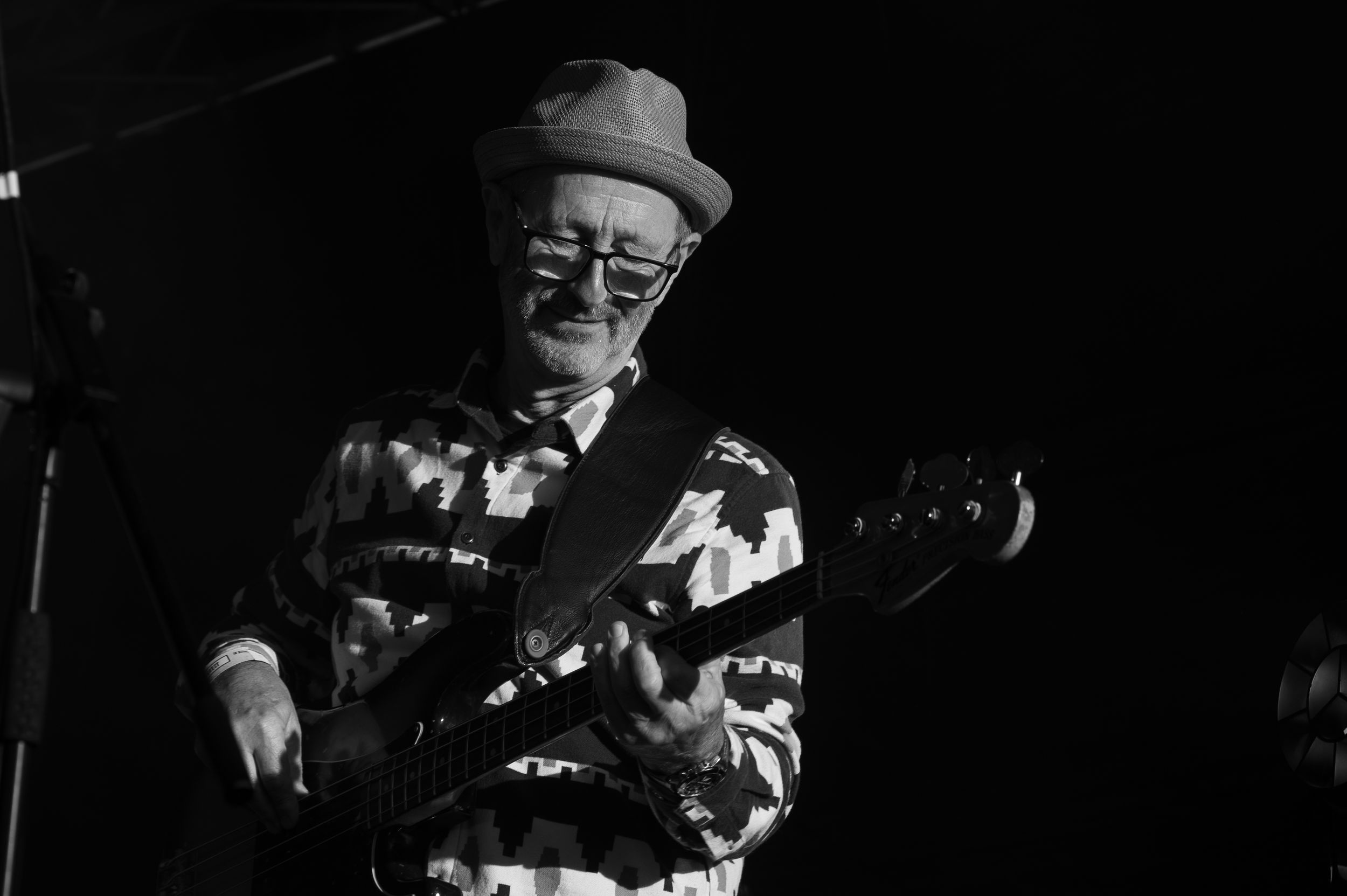 Black and white photo of a musician wearing glasses and a hat playing bass guitar on stage during a performance.