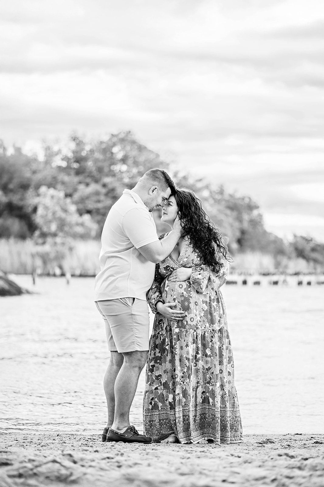 A photograph where a couple lean in together and embrace as mother puts her hands on her belly in a romantic beach maternity session York PA