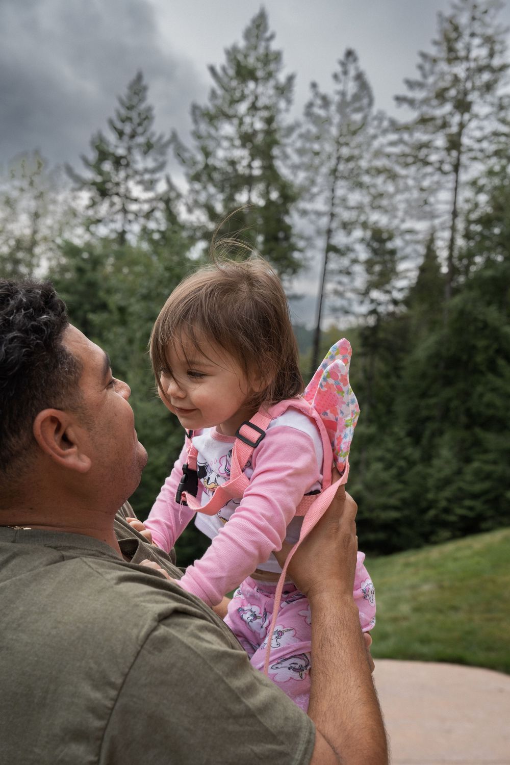 Sweet outdoor moment showing a child in pink fairy wings being lifted up against a backdrop of evergreen trees.