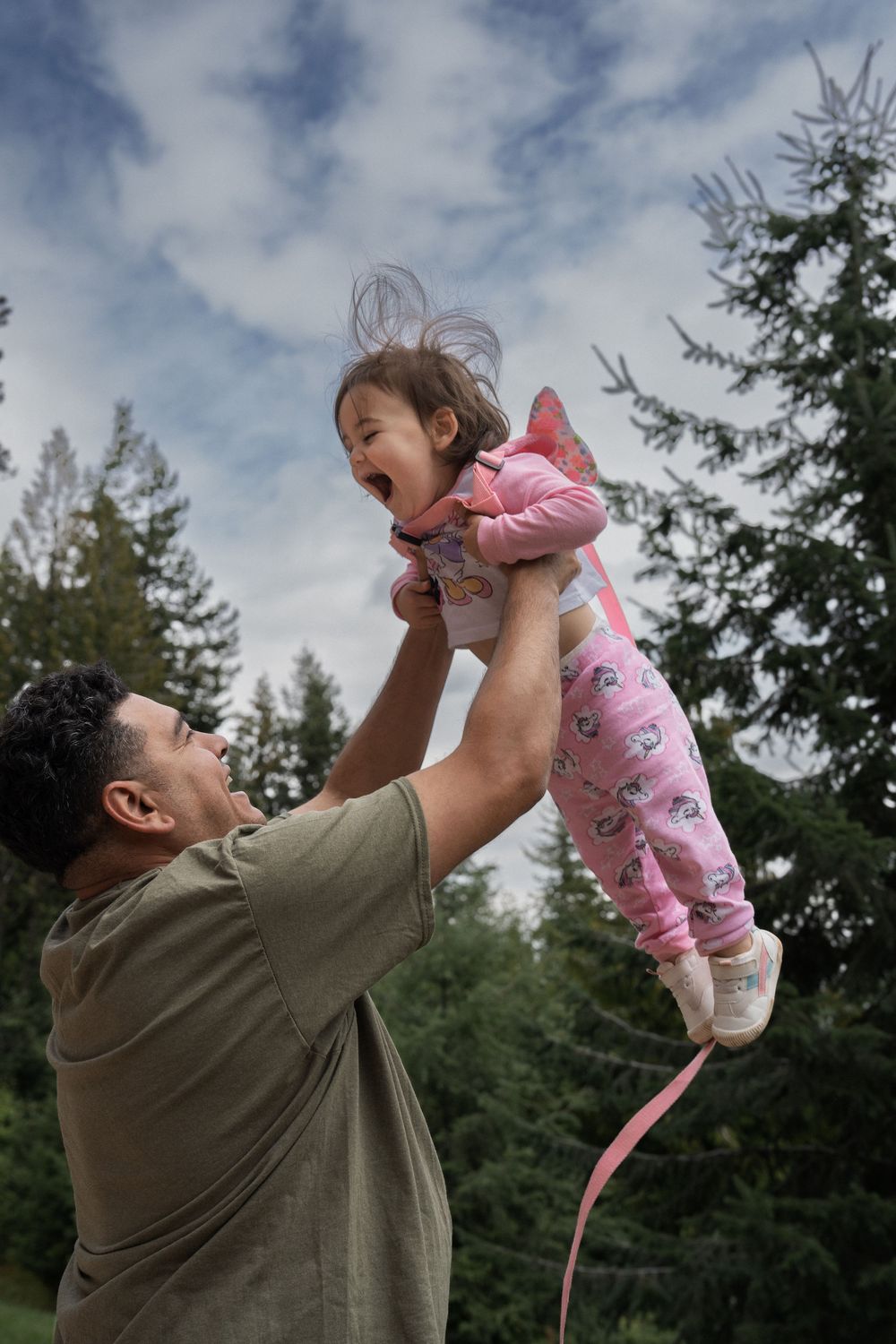 A playful moment outdoors as a child in pink pajamas is lifted high into the air against a cloudy sky.