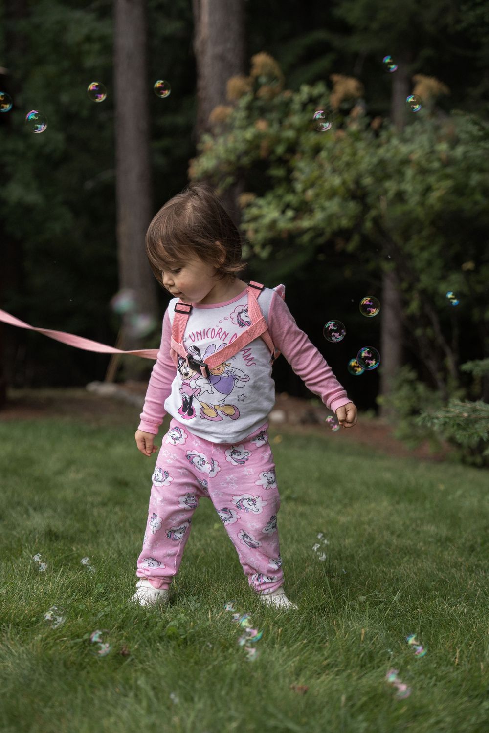 A toddler in pink pajamas chases bubbles while playing on a grassy lawn surrounded by trees.