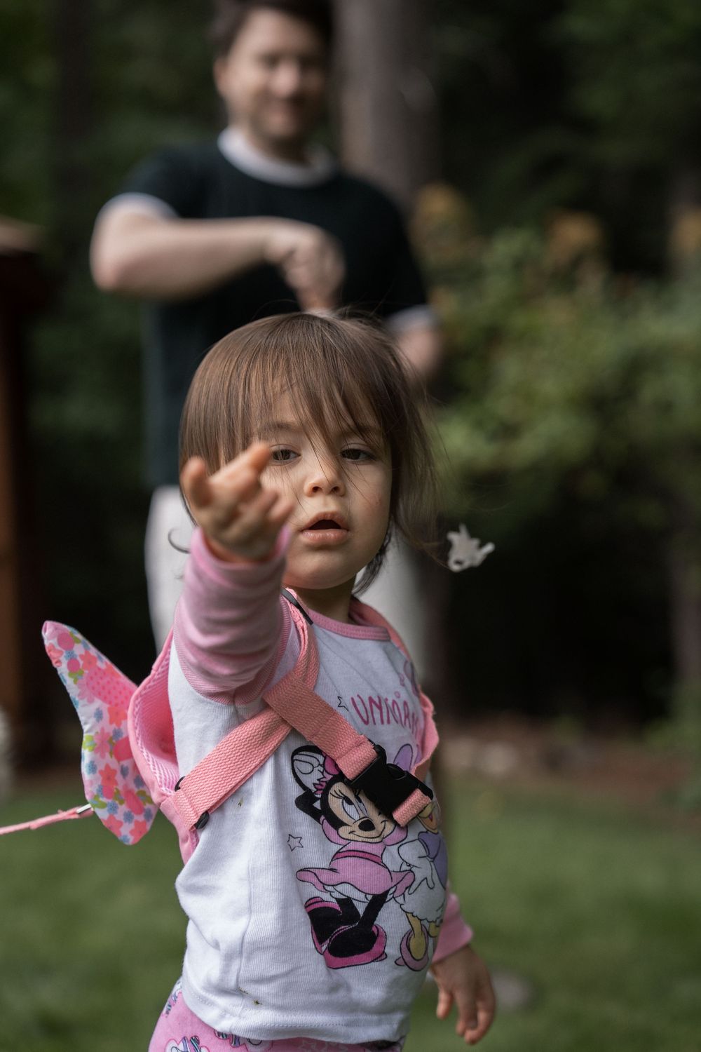 Young child in Minnie Mouse shirt reaches out while playing outdoors with someone in the background.