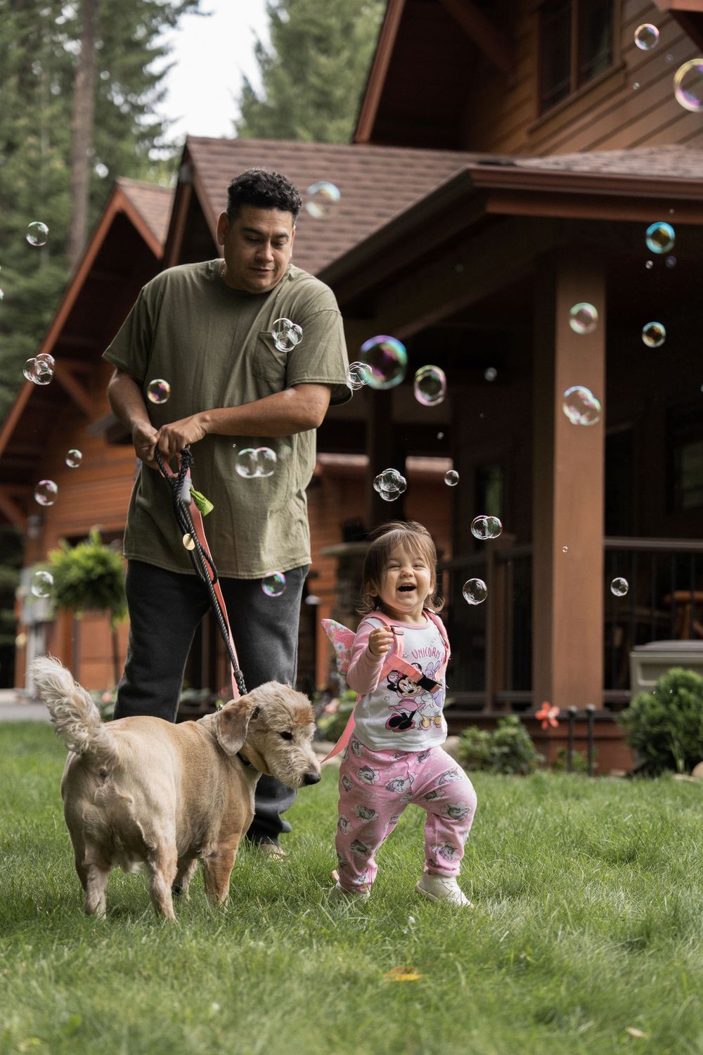 A family and their dog enjoy blowing bubbles together on the lawn in front of wooden cabins.