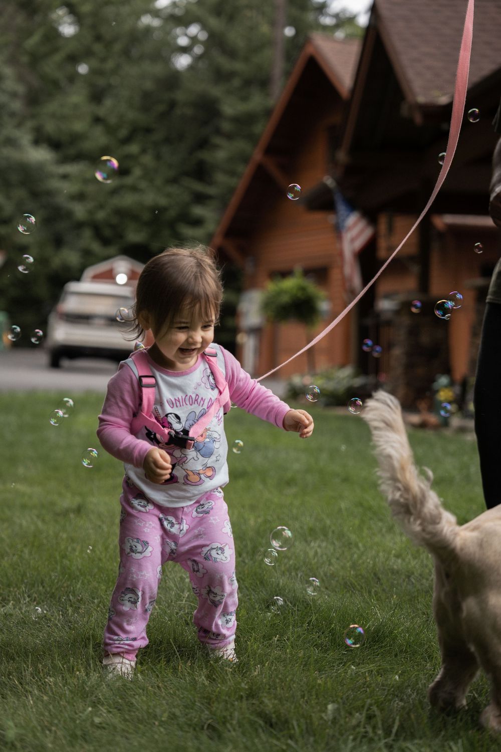 A child in pink pajamas chases bubbles on the grass while playing with a dog.