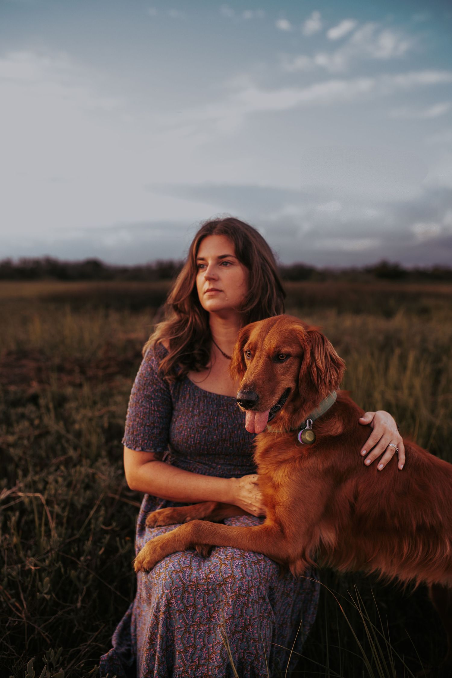 A person in a floral dress sits in a grassy field at sunset with their golden retriever dog.