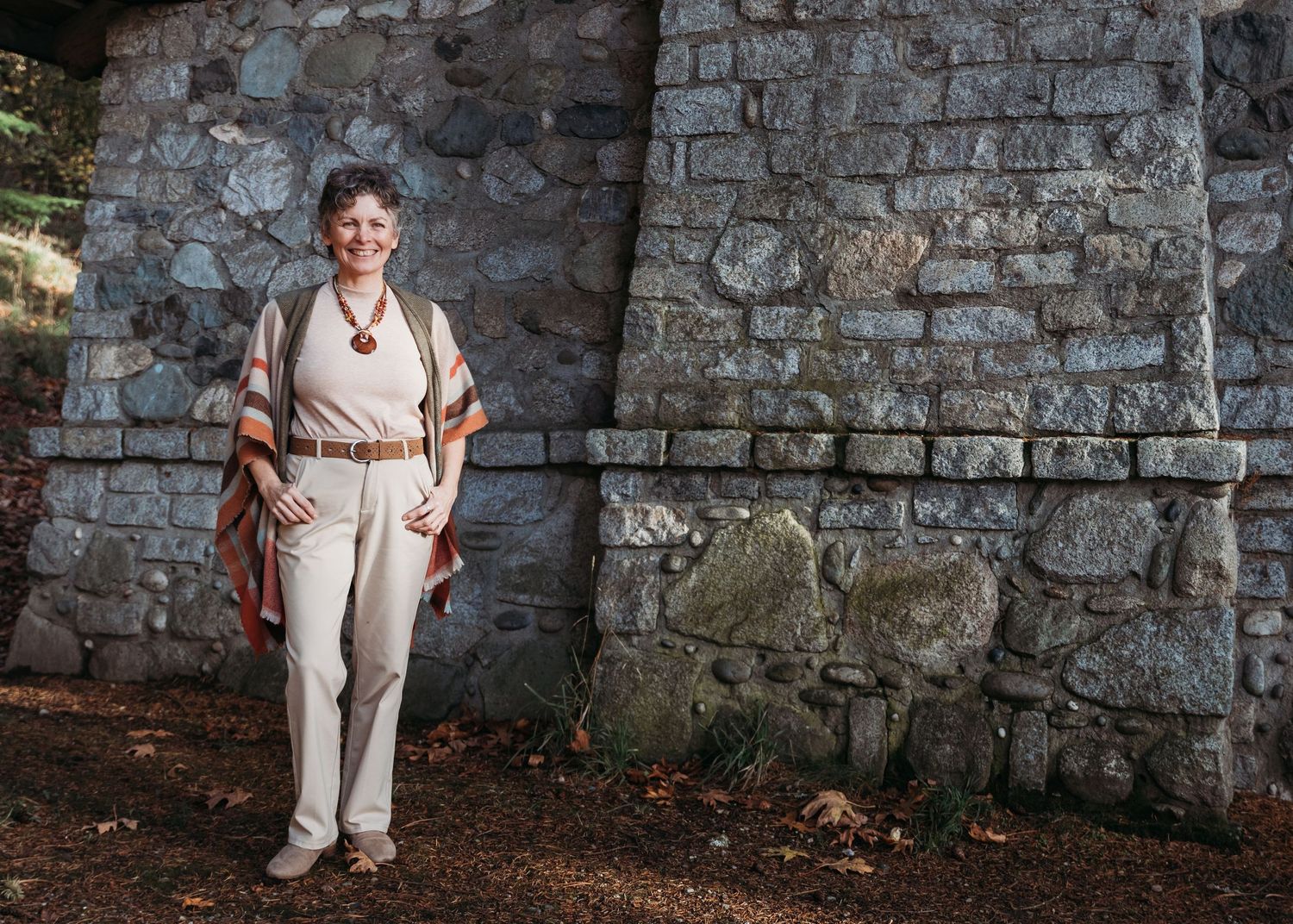 Person in brown blazer and white top smiling while outdoors against blurred natural background.