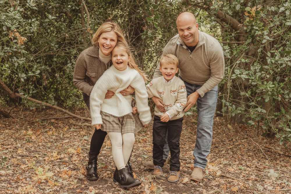 Family in coordinating neutral outfits sits together on fallen tree trunk in autumn forest setting.