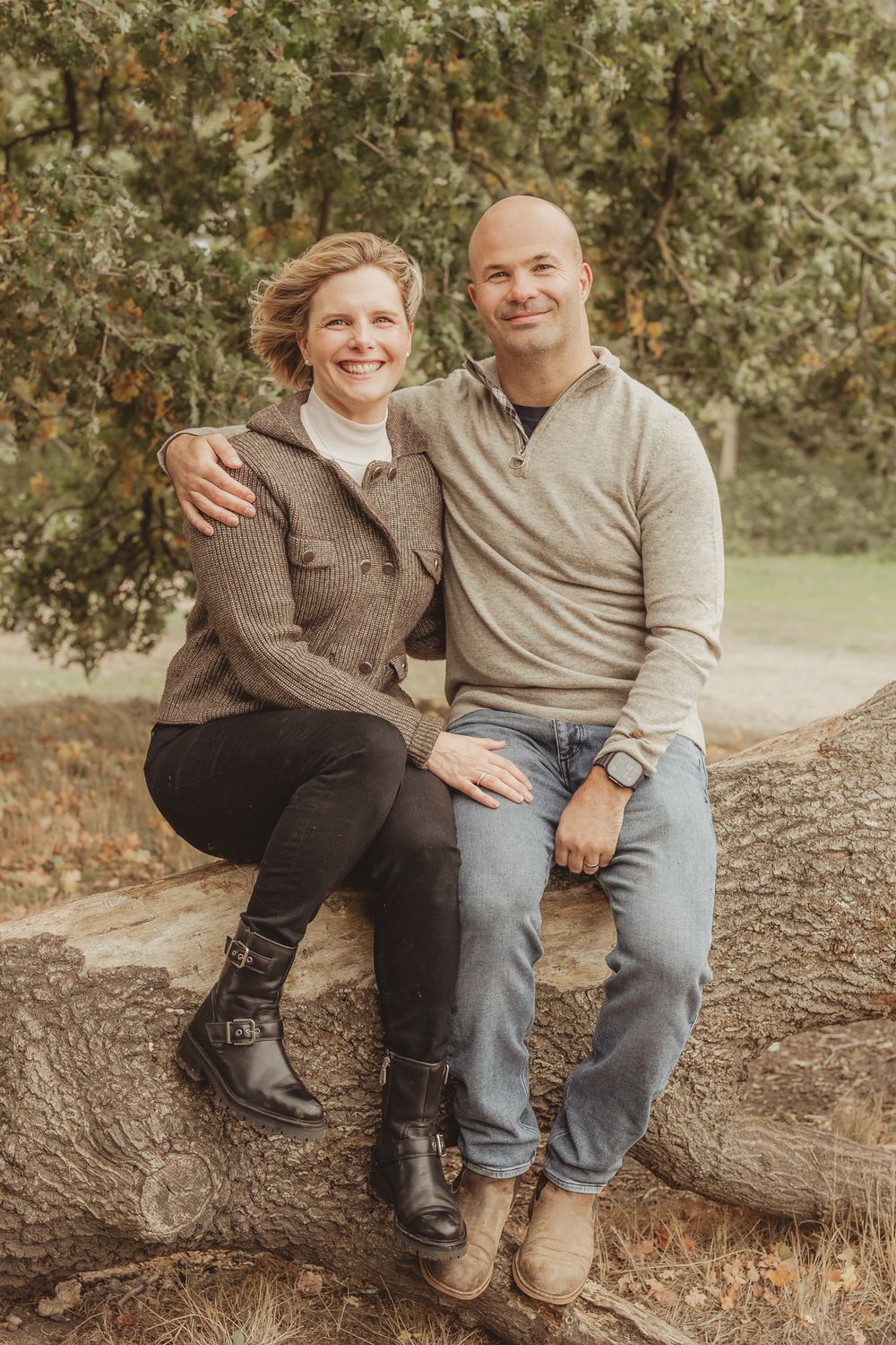 A couple sits together on a fallen tree trunk in an autumn forest setting wearing casual earth-toned clothing.
