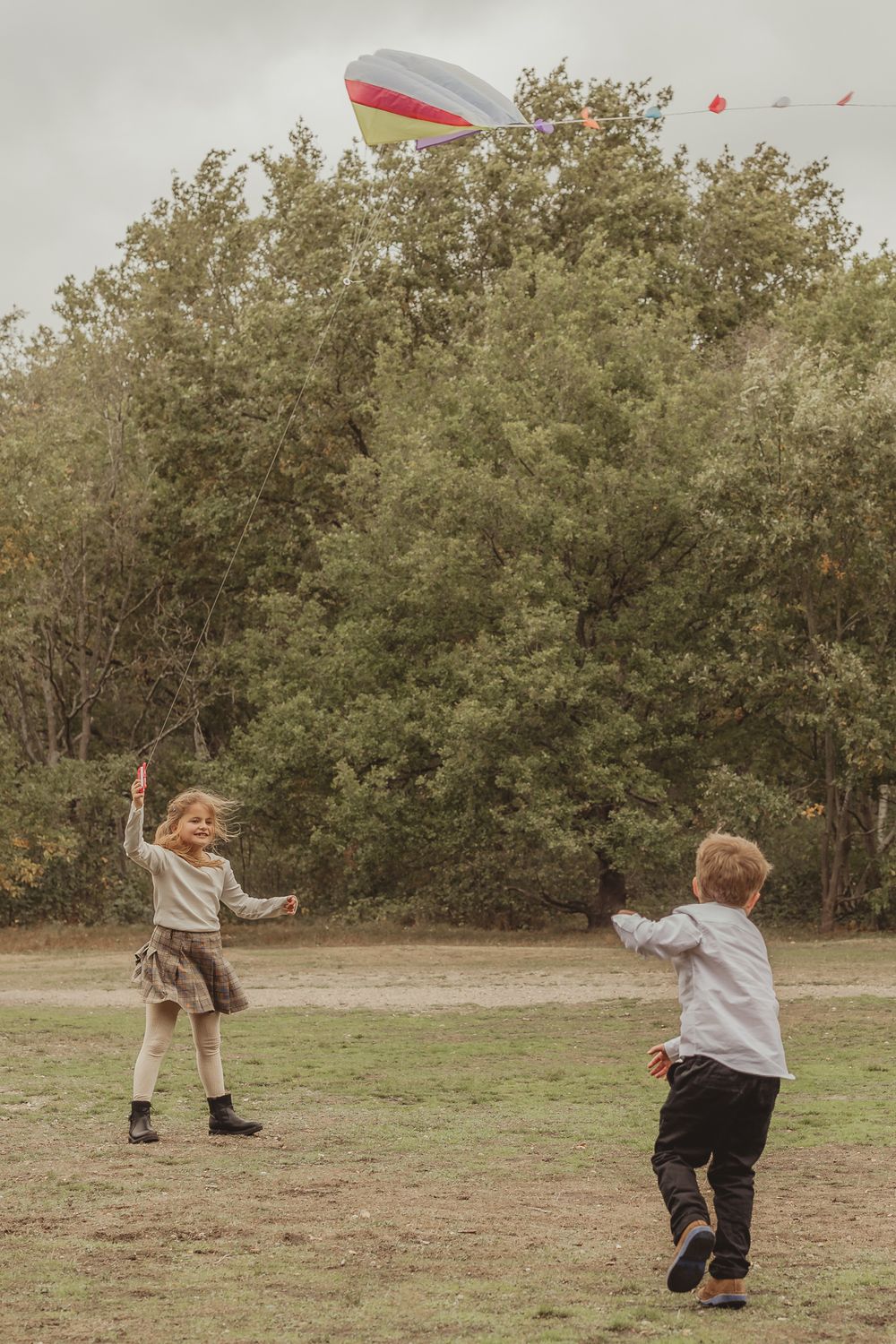 Two children playing with a colorful kite in a grassy field on an overcast day.