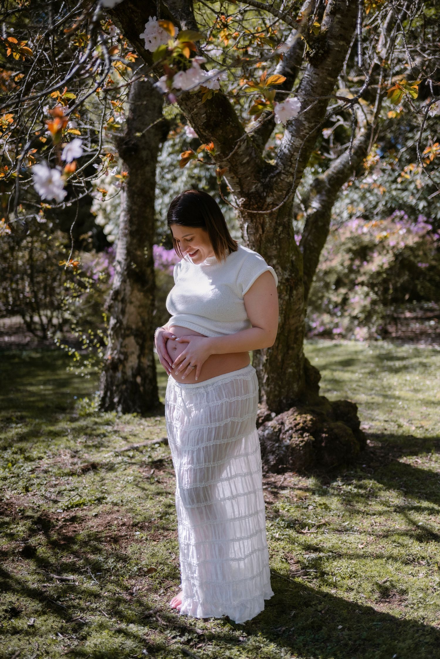 Expectant mother in white tiered dress poses under blooming spring trees.