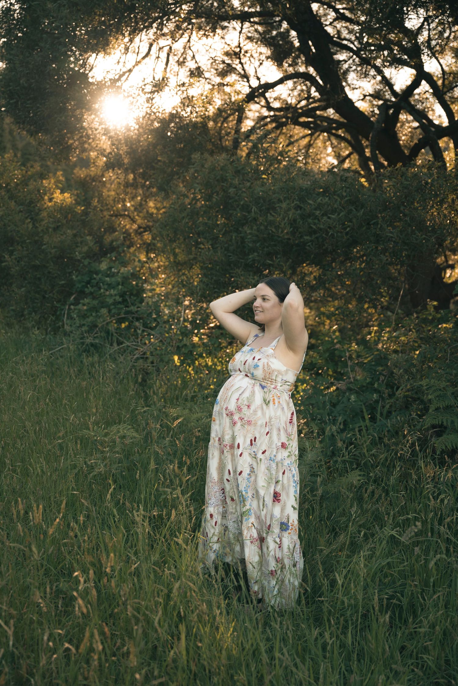 A person in a floral dress stands in tall grass at golden hour as sunlight filters through trees in the background.