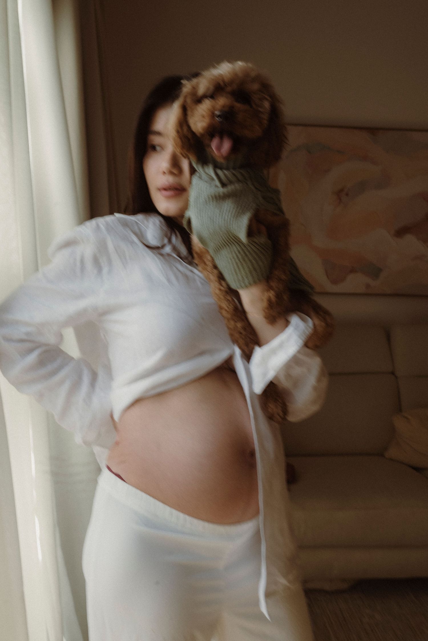 Pregnant woman in white shirt holds a small brown poodle wearing a green sweater, standing by a window.