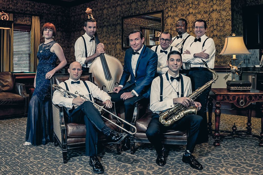 A vintage-styled jazz band poses in elegant tuxedos and a flapper dress in a luxurious room setting.