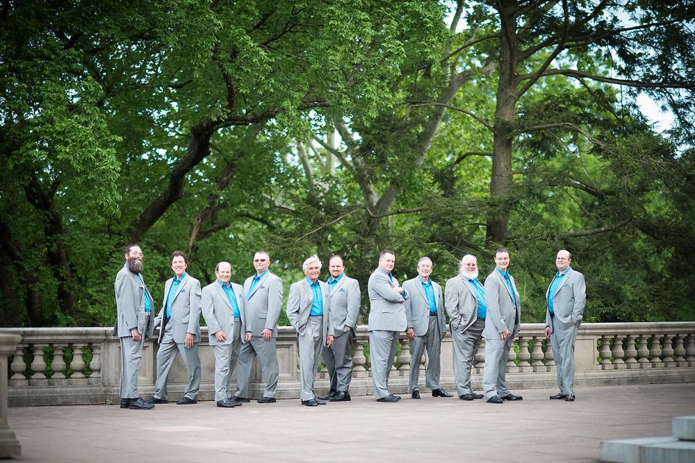 Groomsmen in gray suits with teal ties pose on stone terrace surrounded by lush green trees at outdoor wedding.