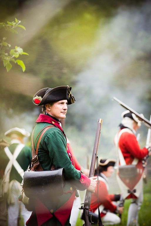 Revolutionary War reenactor in green and red uniform holding musket during historical battle reenactment with smoke.