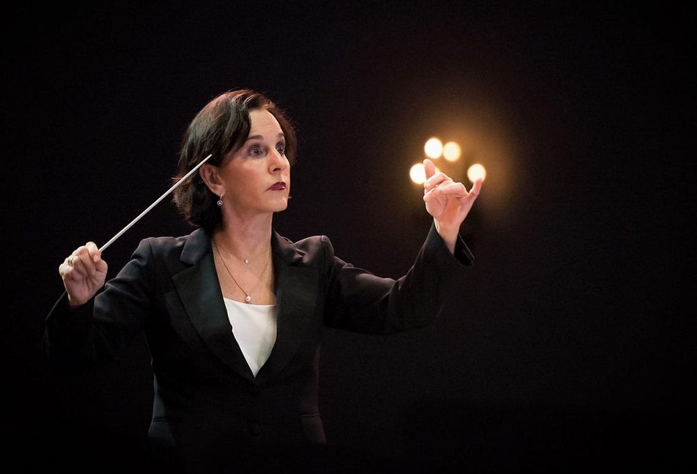 Female orchestra conductor in black blazer holding baton, directing with expressive hands against dark background.