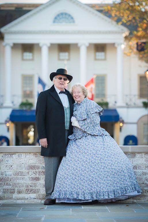 Elderly couple in Victorian period costumes posing in front of a white colonial building with columns.
