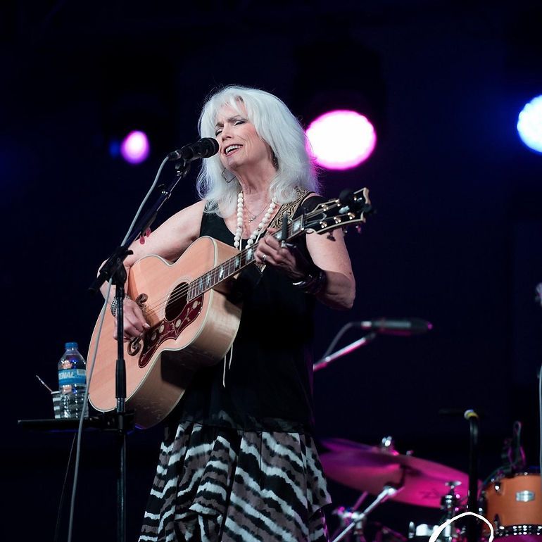 Emmy Lou Harris plays acoustic guitar and sings into a microphone on stage wearing print skirt.