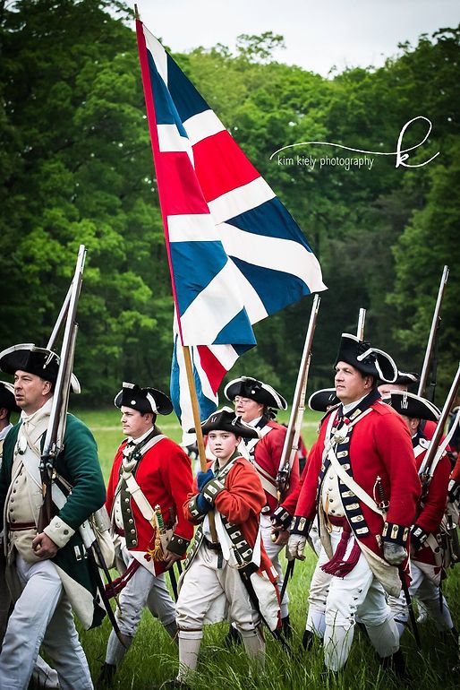 Revolutionary War reenactors in red British uniforms march with a Union Jack flag across a green field.