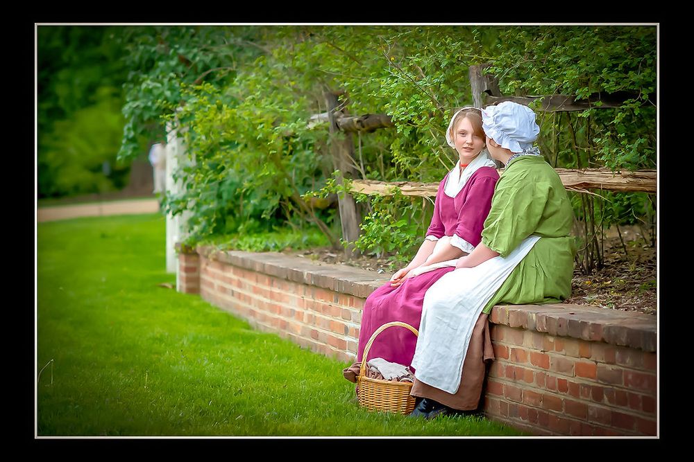 Two women in colonial-era costumes sit on a brick wall in a lush garden setting.