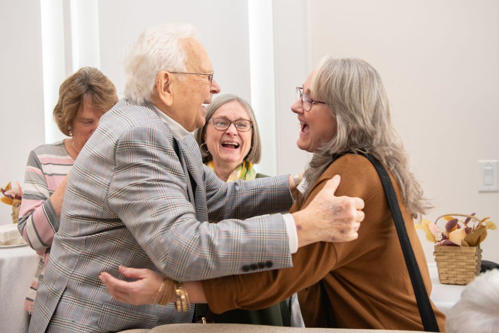 An elderly man warmly embraces a woman while another woman laughs joyfully at a social gathering.Best photographer Williamsburg VA