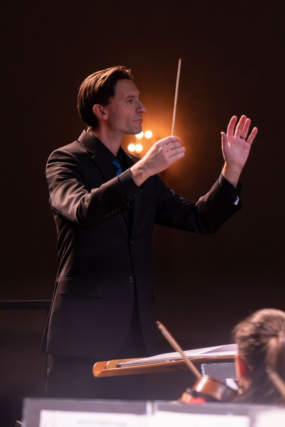 Orchestra conductor in black suit holding baton, leading performance on dimly lit stage with warm lighting Best photographer Williamsburg VA