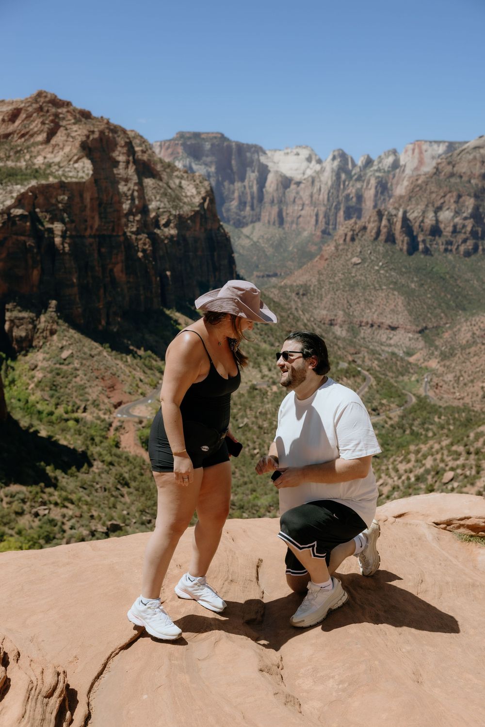 A couple sits together on red rocks overlooking a dramatic canyon landscape on a sunny day.