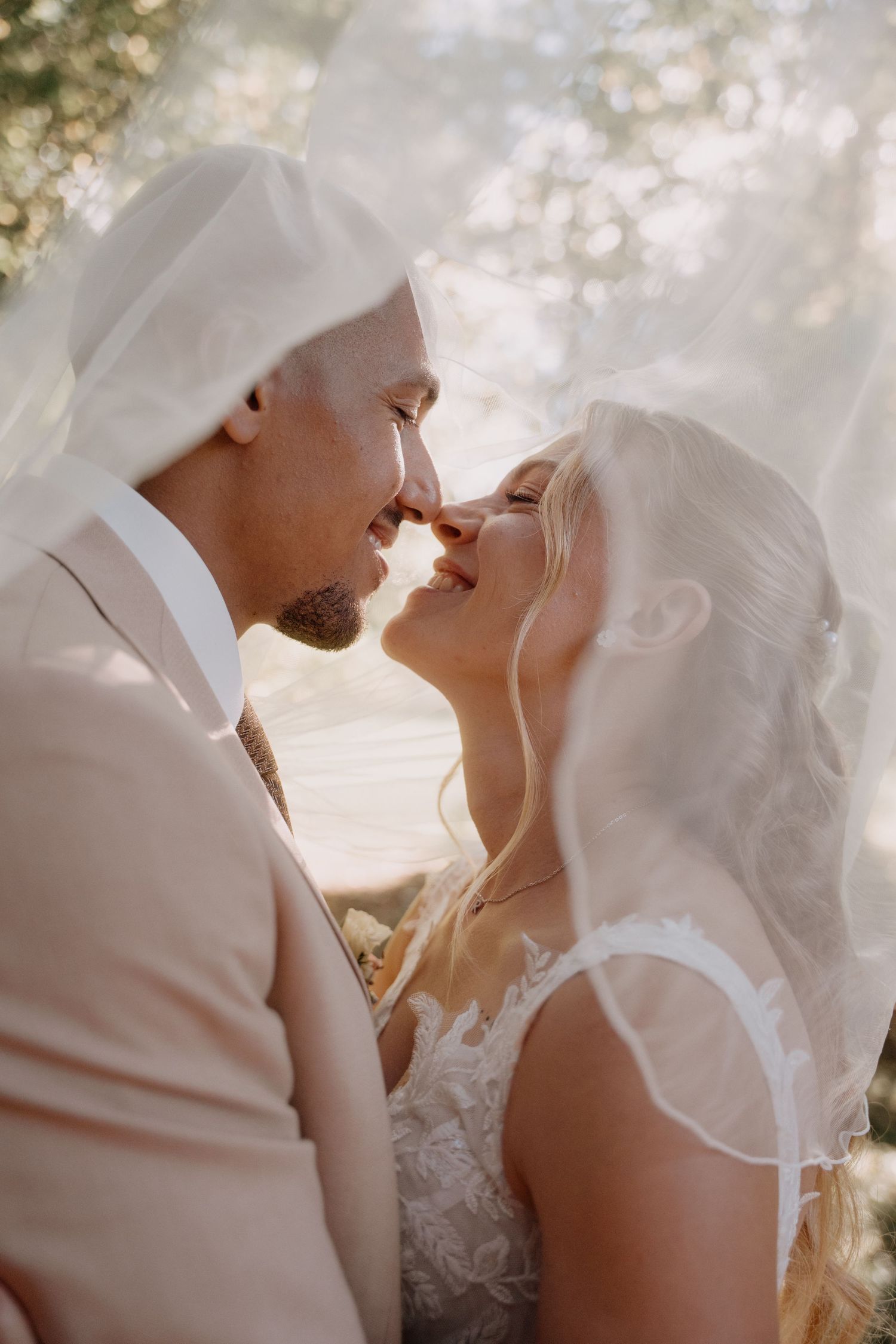 A romantic wedding couple shares an intimate moment under a flowing white veil in soft natural light.