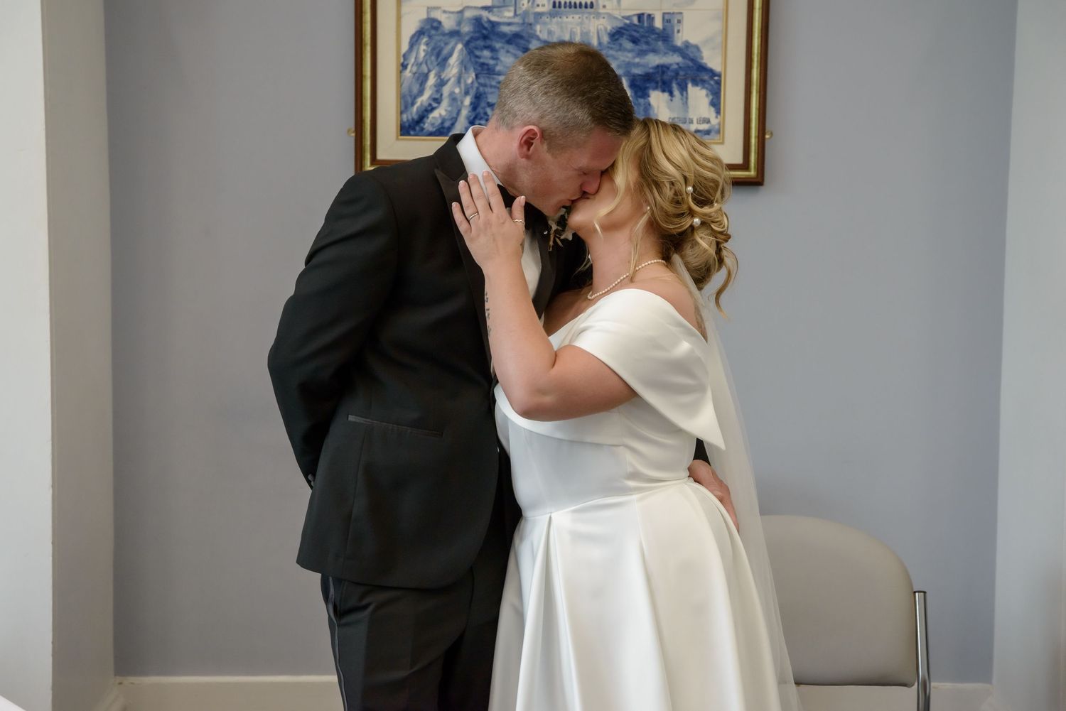 A couple in wedding attire share a romantic kiss in front of a gray wall with framed artwork.