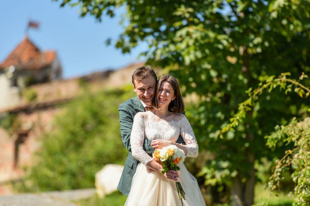 A couple embraces outdoors on their wedding day with green foliage and a castle in the background.