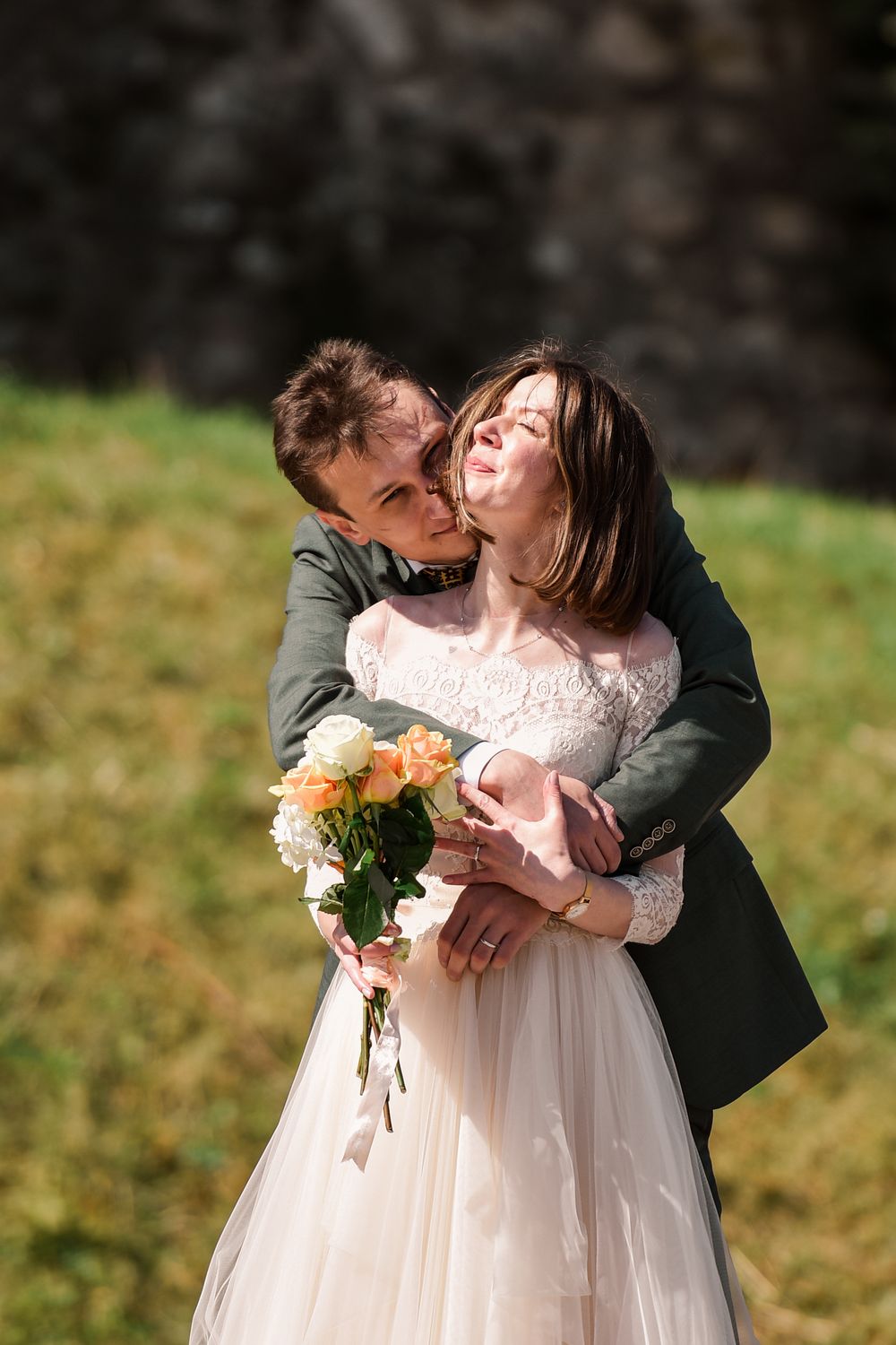 A romantic wedding couple embraces in a sunlit outdoor setting with the bride holding a bouquet of flowers.