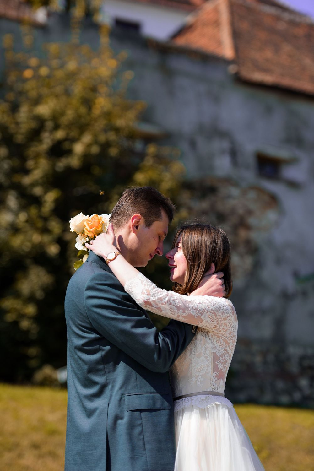 A romantic wedding couple embraces outdoors in front of a historic stone building on a sunny autumn day.