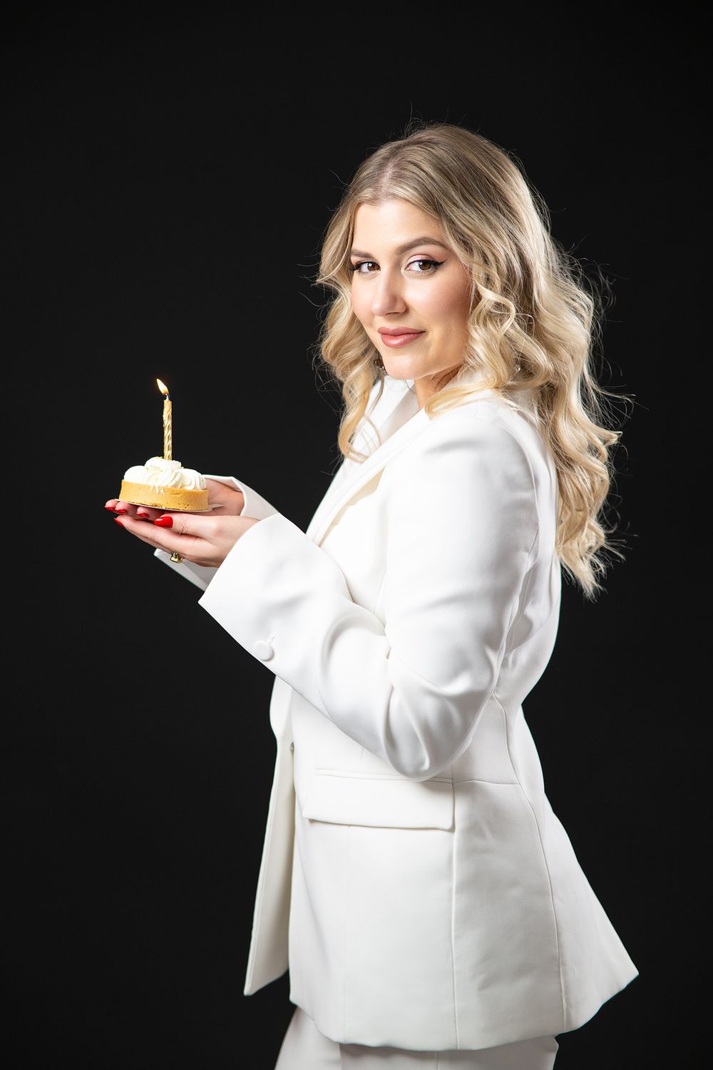 A person in a white blazer poses against a black background holding a birthday cupcake with a lit candle.