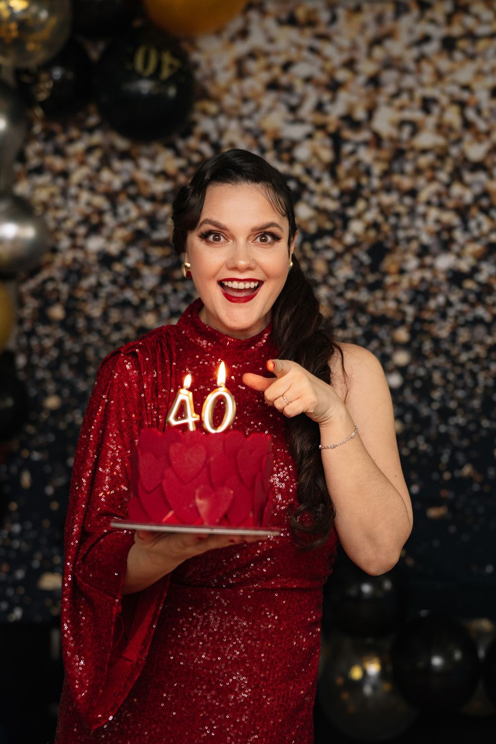 A person in a sparkly red dress poses with a birthday cake featuring lit number 40 candles against a bokeh background.