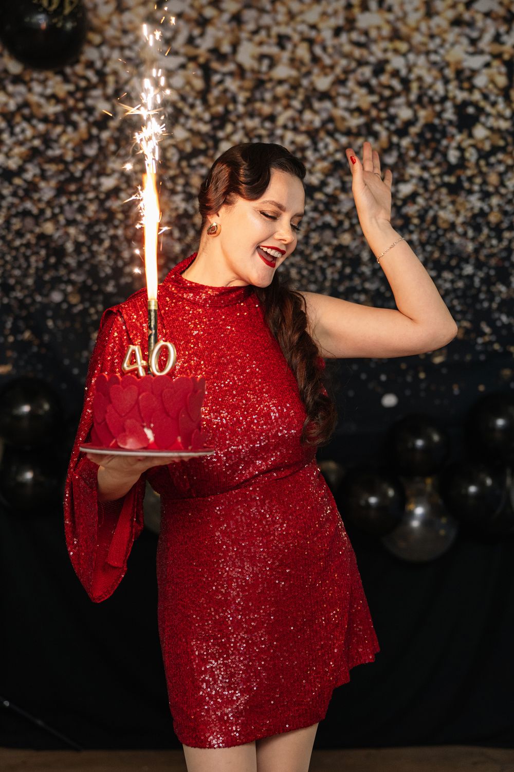 A person in a sparkling red dress dances joyfully while holding a sparkler against a dark glittering background.