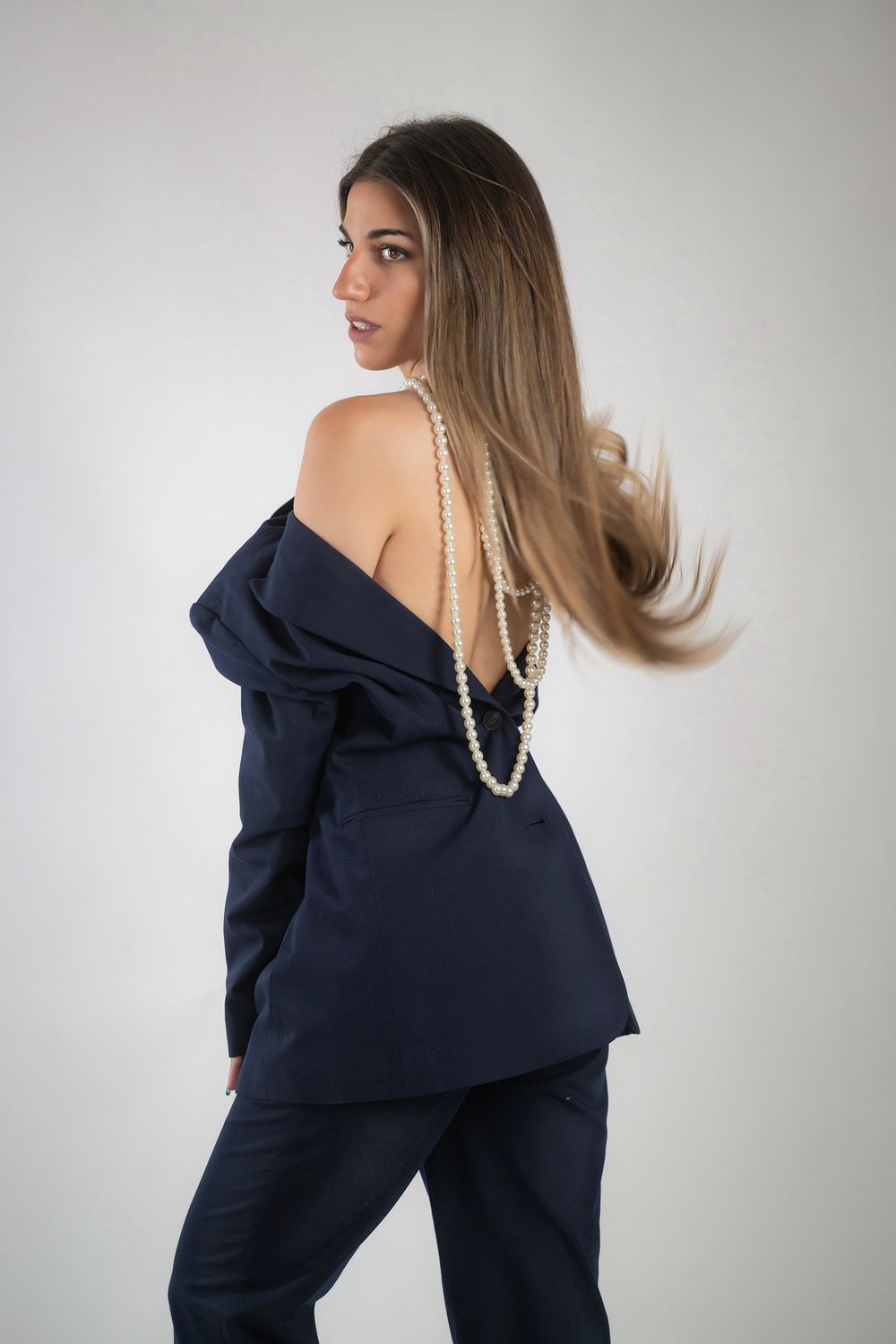 A model wearing a navy blue one-shoulder dress with dramatic ruffled neckline and pearl necklace poses in a studio setting.