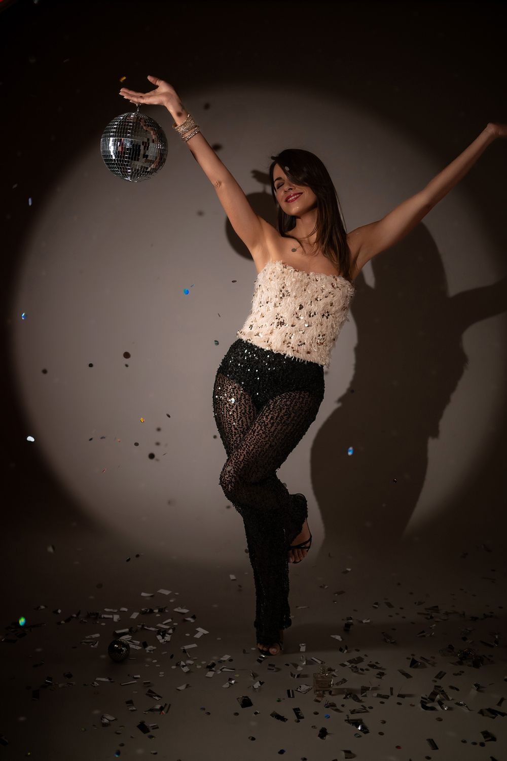 A person in a sparkly dress dances joyfully with confetti falling in a dimly lit studio setting.