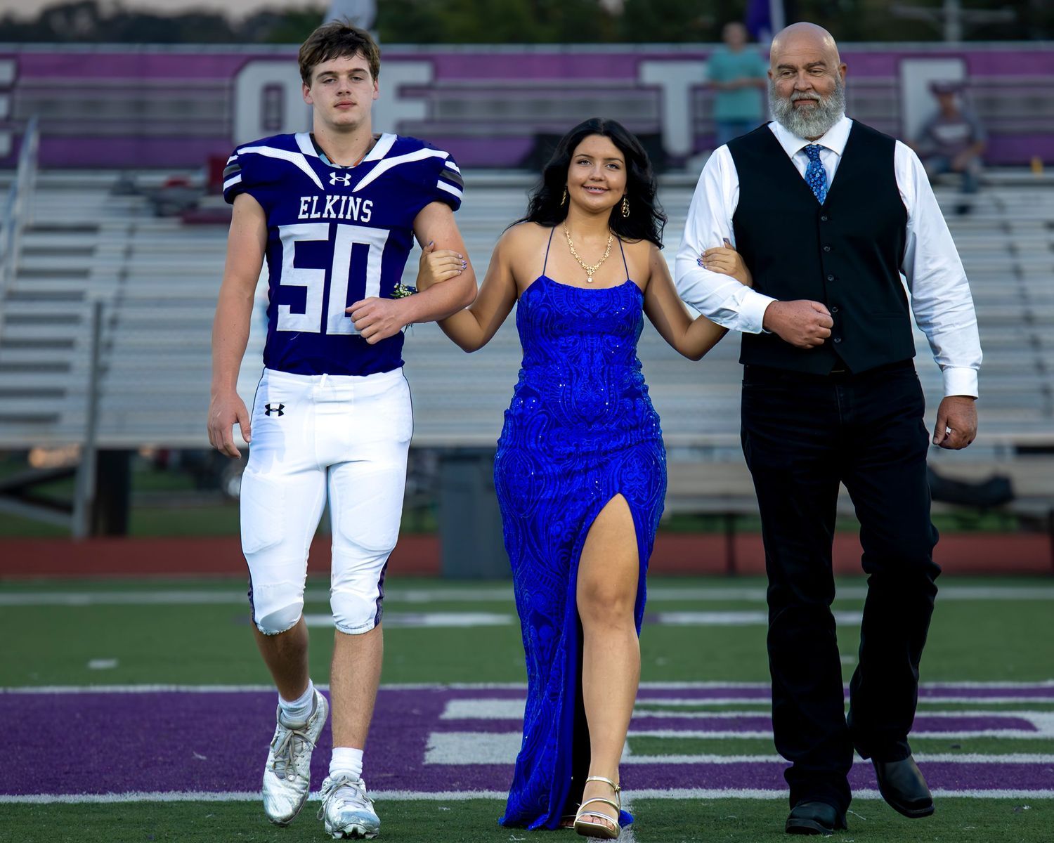 Football players and attendants walk onto field during homecoming ceremony in formal attire.