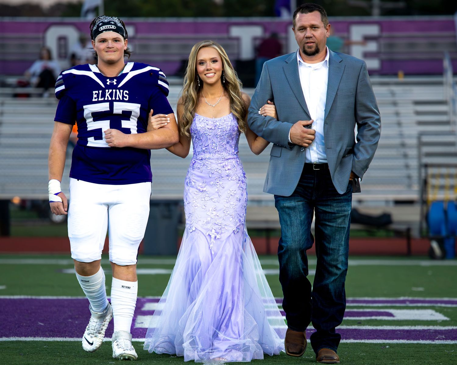 A group walks across a football field with someone wearing a lavender formal dress and others in casual attire.