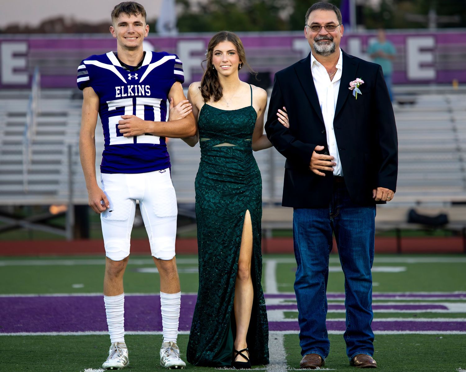 Football player in white uniform poses with couple in formal attire on sports field during photo session.