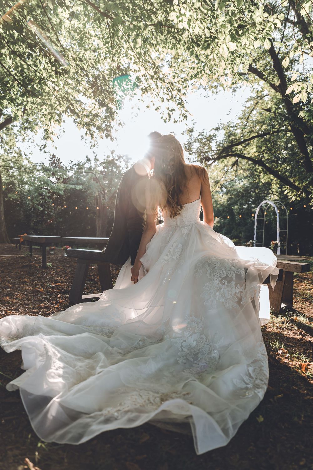 A wedding dress flows elegantly in sunlit forest setting as sunbeams filter through tree branches.