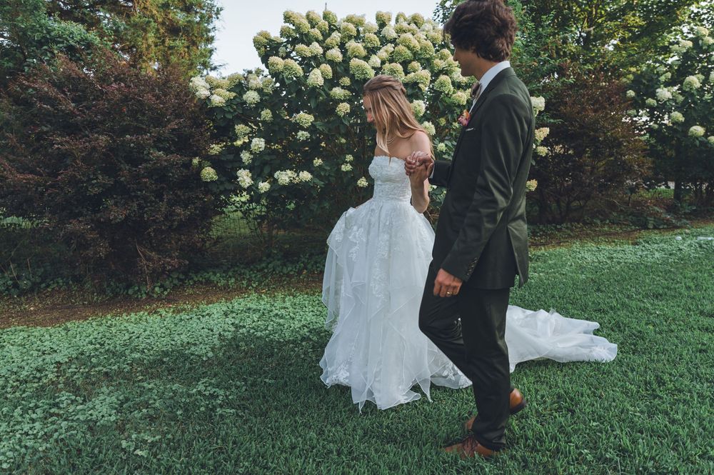 A couple in wedding attire embraces on a green lawn with blooming trees in the background.
