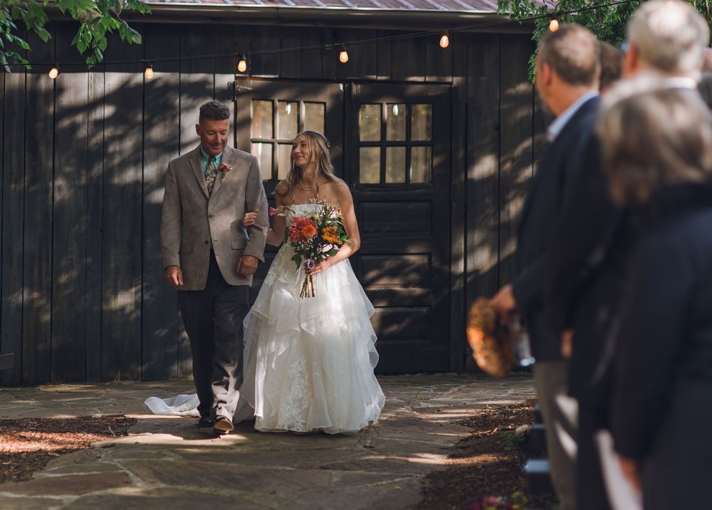 A wedding couple walks down a tree-lined path as guests look on during an outdoor ceremony.