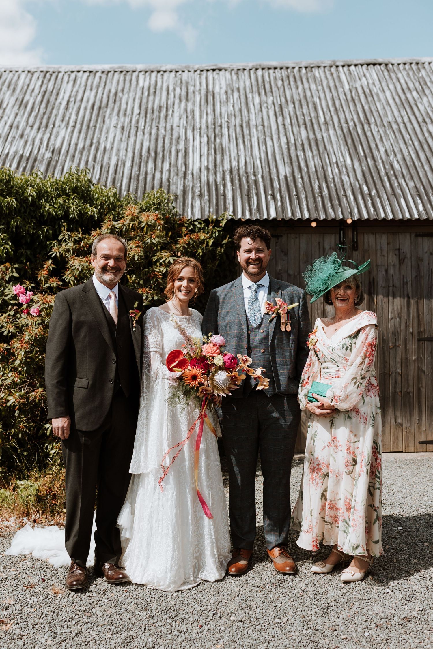 A group in formal wedding attire poses together against a rustic wooden wall outdoors.